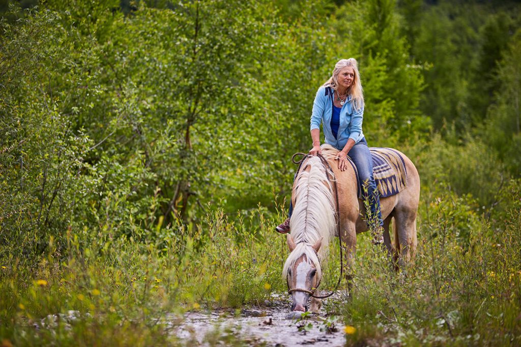 Das Foto zeigt Tina Boche ohne Sattel ihrem Haflinger Paco sitzend. Paco trinkt an einem Bach mitten in der Natur.